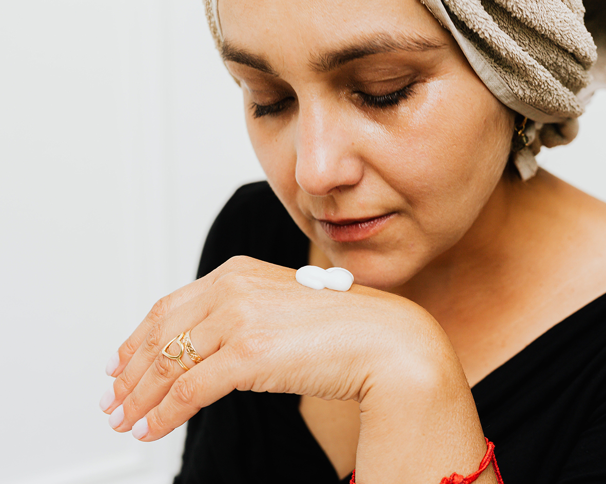 Una donna ha della crema viso invernale sulla mano e si avvicina per poterla applicare sulla pelle. Si trova in bagno e ha un asciugamano in testa.